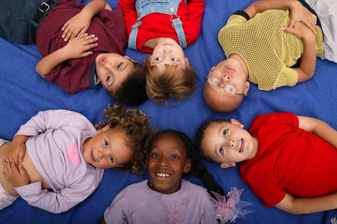 Six young children of diverse backgrounds lying in a circle on a blue mat with their heads together, smiling up at the camera.