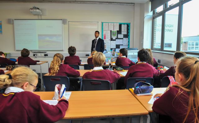A teacher stands at the front of a classroom presenting a video on an interactive whiteboard to pupils in burgundy school uniforms seated at desks.