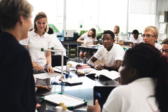 A teacher talks with secondary school pupils in white uniforms gathered around a lab table during a science lesson.
